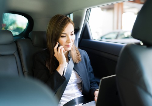 A young business woman in the back seat of a car works on her laptop and listens on her phone after searching for a "Taxi Near Me"